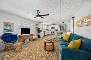 A modern living room with a television, seating, a kitchen in the background, and a ceiling fan.