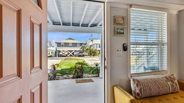 A room with a wooden door, a yellow sofa, a window with blinds, and a view of a street and houses outside. - Stay Longer, Save More (Fernandina Beach)