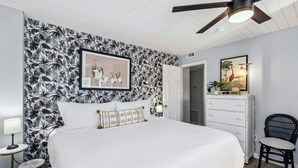 A bedroom with a large bed, a ceiling fan, a dresser, a chair, and a door leading to another room. - Stay Longer, Save More (Fernandina Beach)
