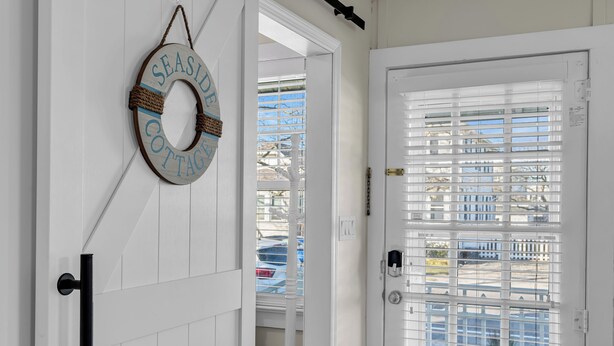 Une porte blanche avec une fenêtre ornée de stores horizontaux, un carillon et une plaque fixée au mur portant l’inscription « SEASIDE COTTAGE ».