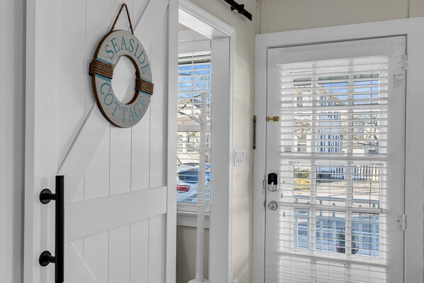 Una puerta blanca con una ventana que tiene persianas horizontales, un timbre y un letrero en la pared que dice 'SEASIDE COTTAGE'.