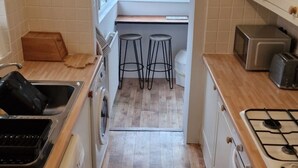Cocina compacta con lavadora, estufa y lavamanos. Hay taburetes cerca de la ventana y un reloj en la pared.