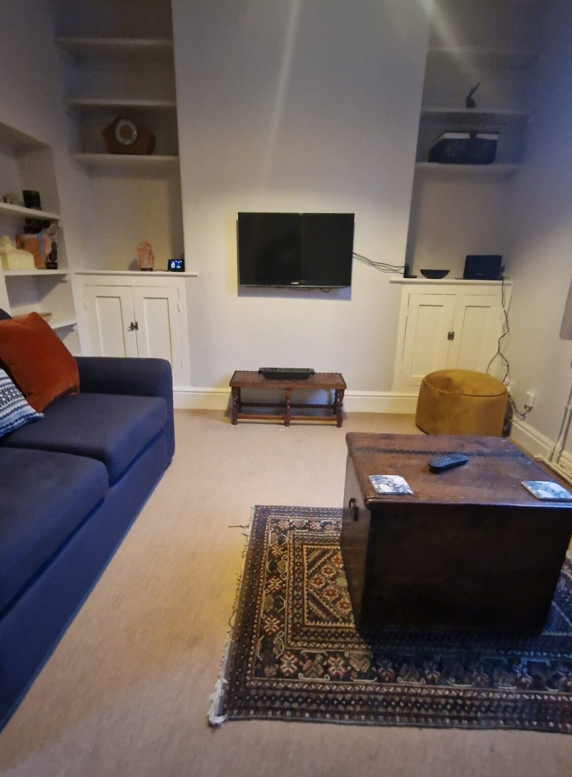Una sala de estar con un sofá azul, una mesa de centro de madera, un tapete estampado, un televisor en la pared y un mueble con estantes.