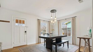 Ein Essbereich mit einem schwarzen Tisch und einer Bank, einer Chandelüster und Blick ins Freie durch große Fenster.