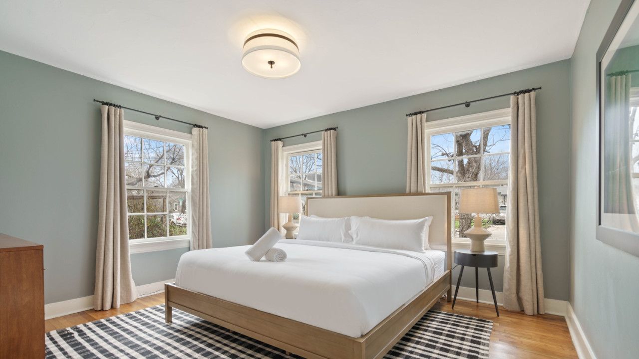 A bedroom with a large bed, two windows with curtains, a bedside table with a lamp, and a checkered rug.