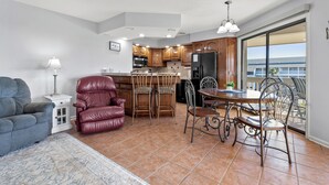 A modern kitchen with wooden cabinets, a dining area with a table and chairs, a red leather chair, and a view of a building through a glass door. - Tilghman Lakes By the Sea (North Myrtle Beach)