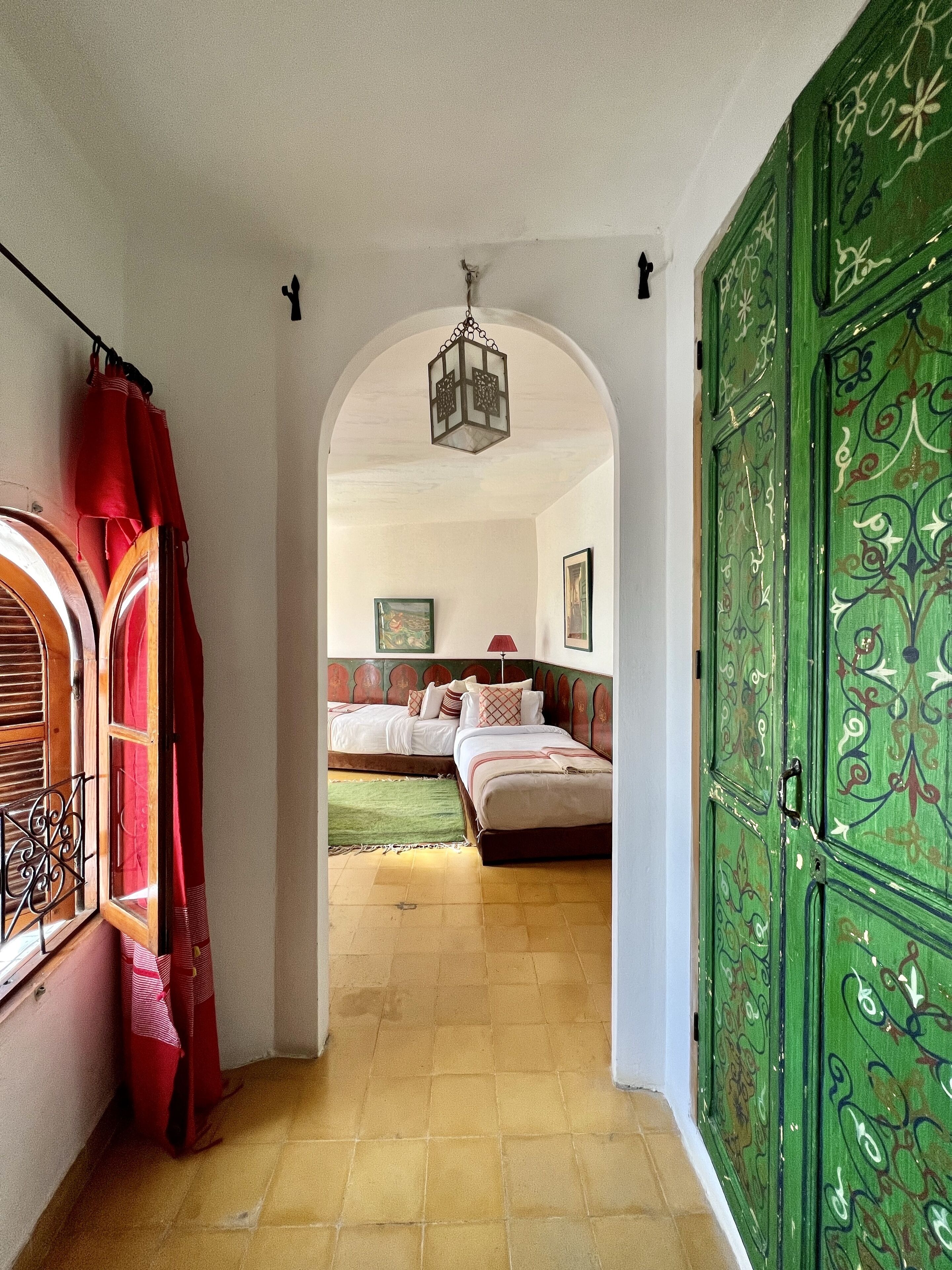 Un pasillo estrecho que conduce a un dormitorio con una cama, una puerta verde con detalles intrincados y un farol colgante.