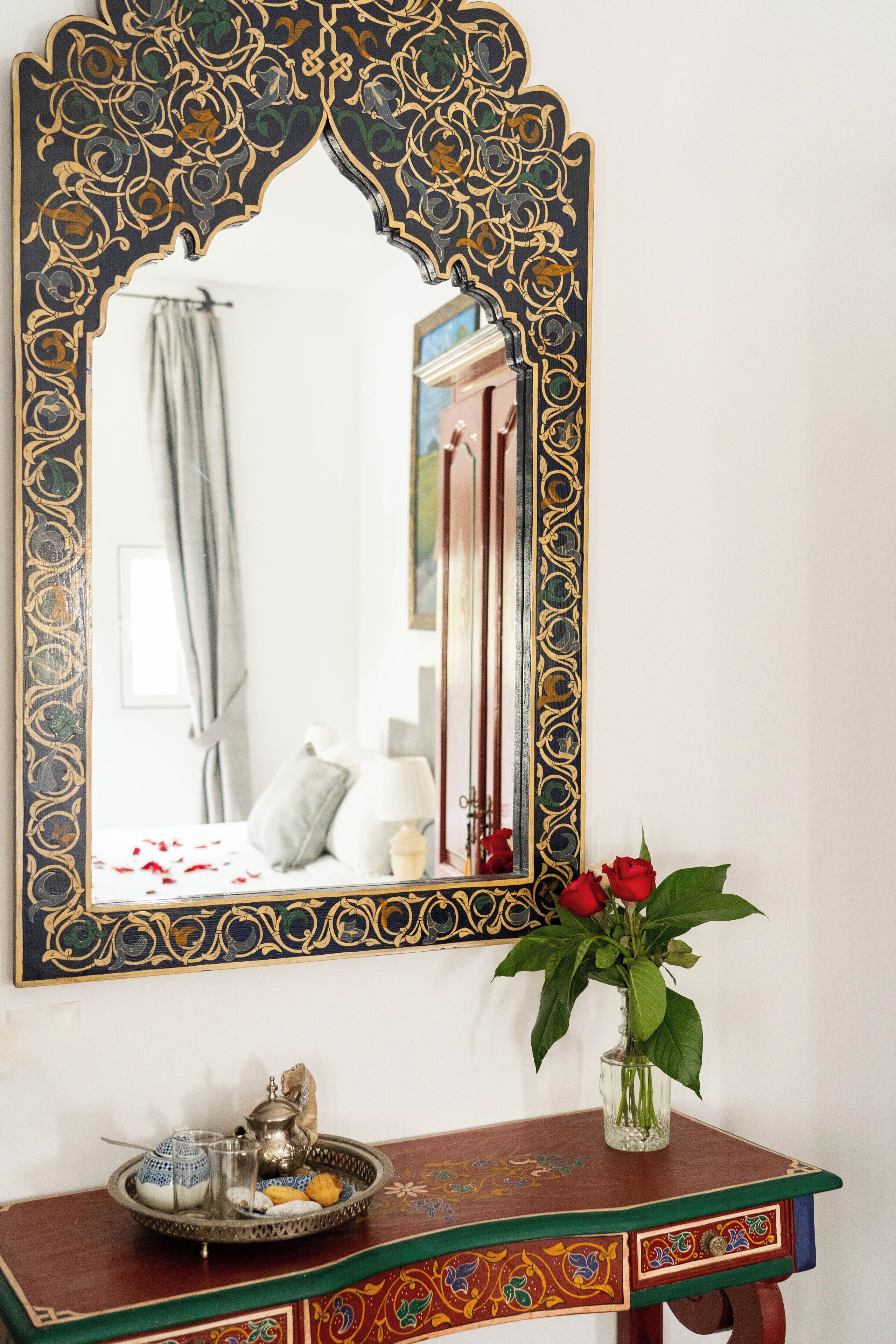 Un miroir avec un cadre noir et doré orné, une table en bois sur laquelle se trouve un plateau rempli d’objets, et un vase contenant des roses rouges.
