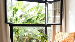 Una habitación con un ventanal amplio, una silla de fibras naturales, una mesa de madera con una bandeja y un suelo de baldosas.