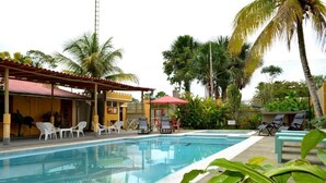 A swimming pool with lounge chairs and umbrellas, surrounded by palm trees and a building with a red roof.