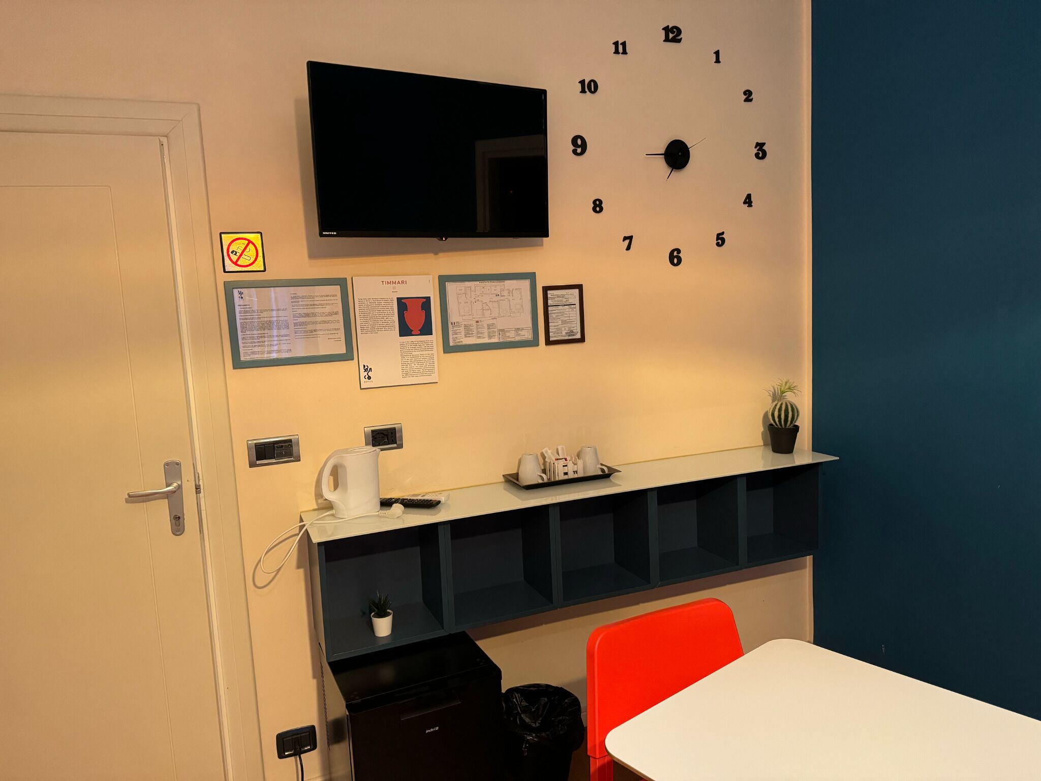 A room with a white door, a black clock, a flat-screen TV, a white table, a red chair, a small potted plant, and a trash bin.