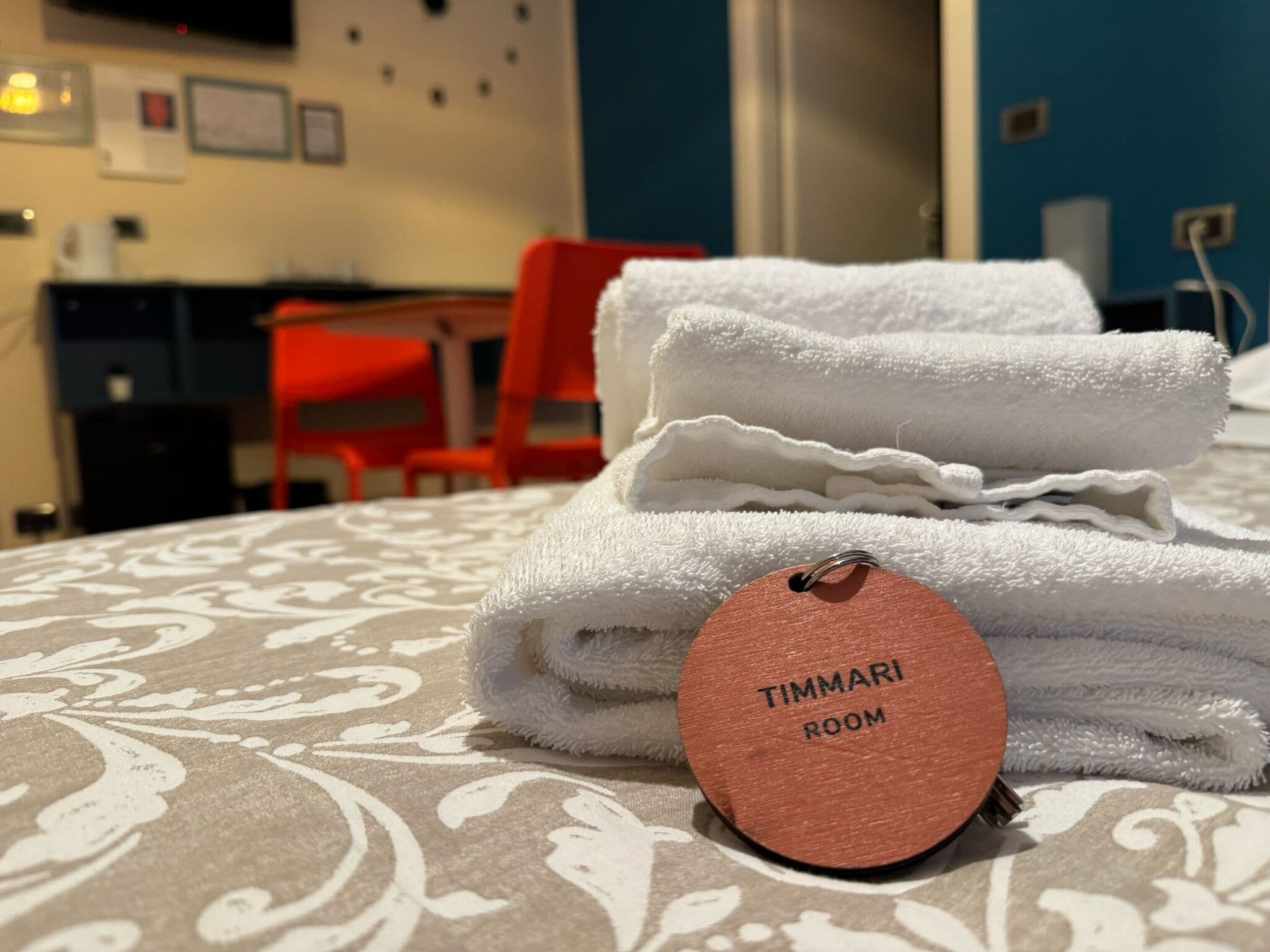 A neatly folded white towel with a tag labeled "TIMMARI ROOM" on a patterned bedspread.
