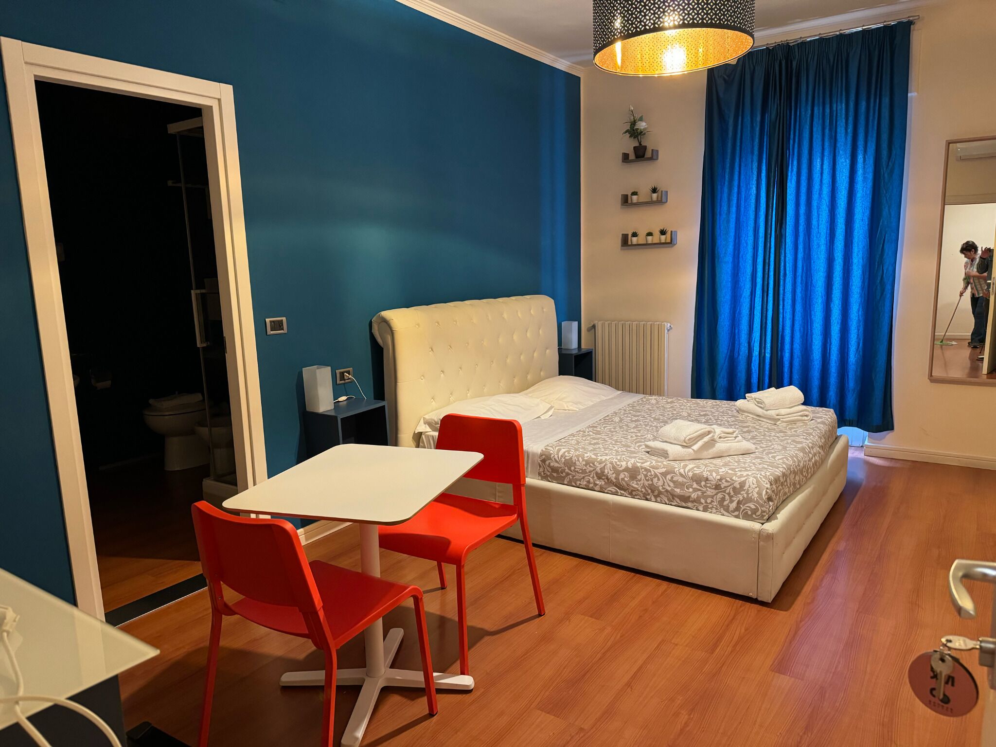 Un dormitorio con cama, escritorio y silla. Hay una ventana con cortinas, un radiador y una estantería con plantas.
