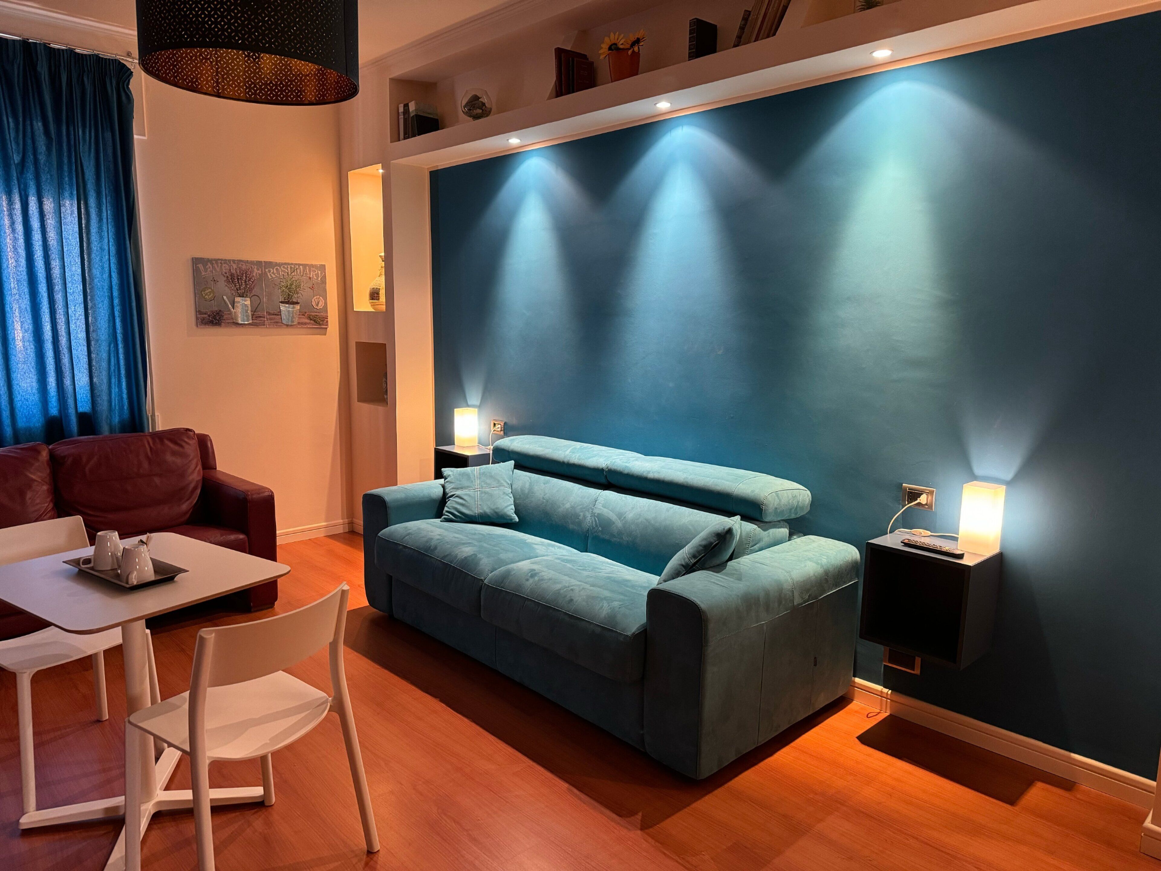 A modern living room with a blue sofa, a white table, and a red armchair. There are two wall-mounted lamps and a ceiling light.