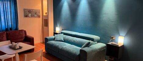 A modern living room with a blue sofa, a white table, and a red armchair. There are two wall-mounted lamps and a ceiling light.