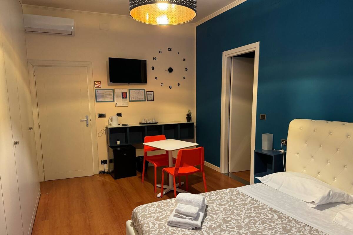 Een slaapkamer met een bed, een bureau met stoel, een klok en een televisie aan de muur.