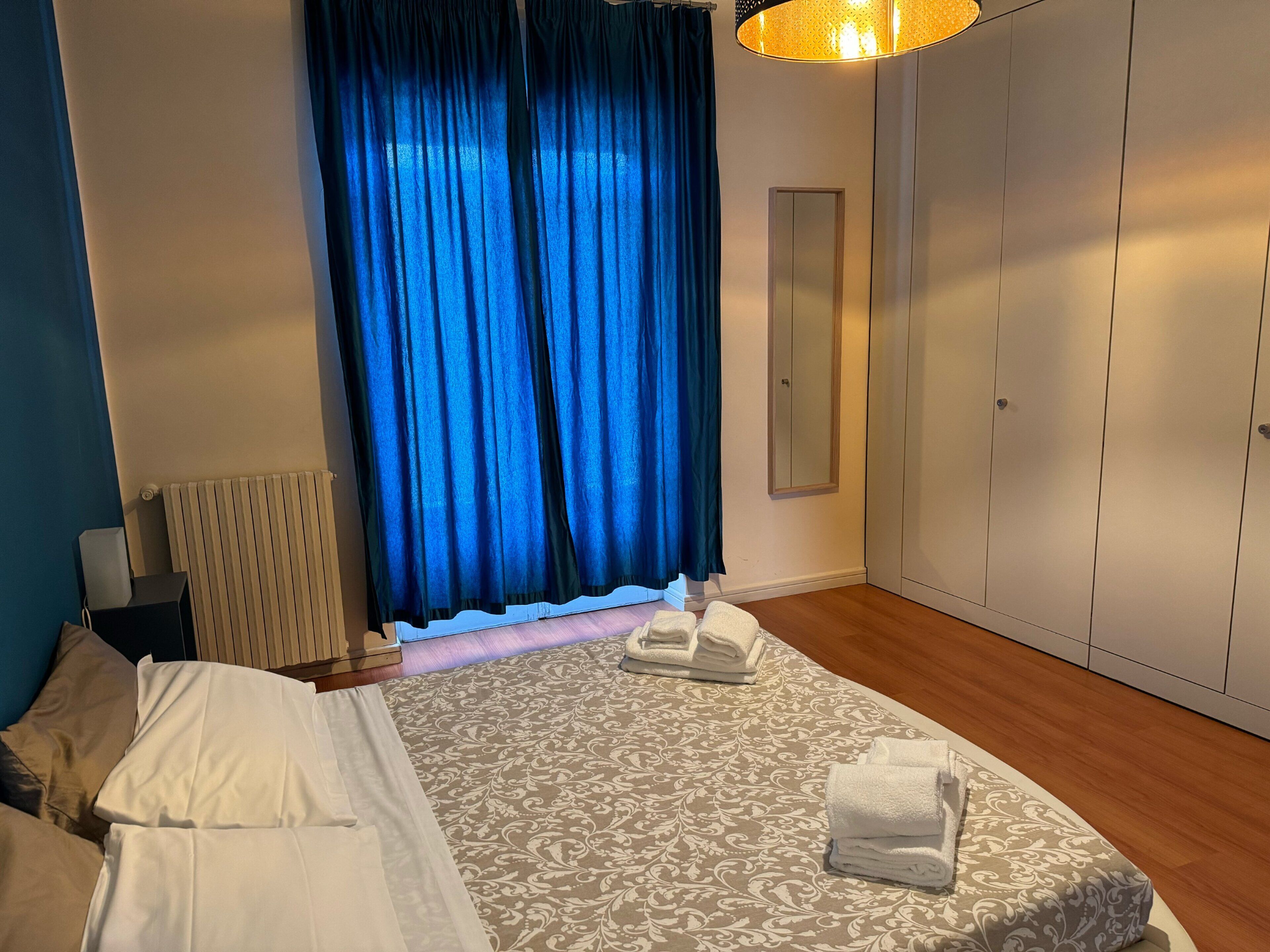 Un dormitorio con cama, mesita de noche, espejo, armario y ventana con cortinas azules.