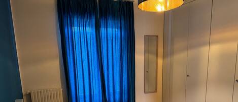 A bedroom with a bed, a bedside table, a mirror, a wardrobe, and a window with blue curtains.
