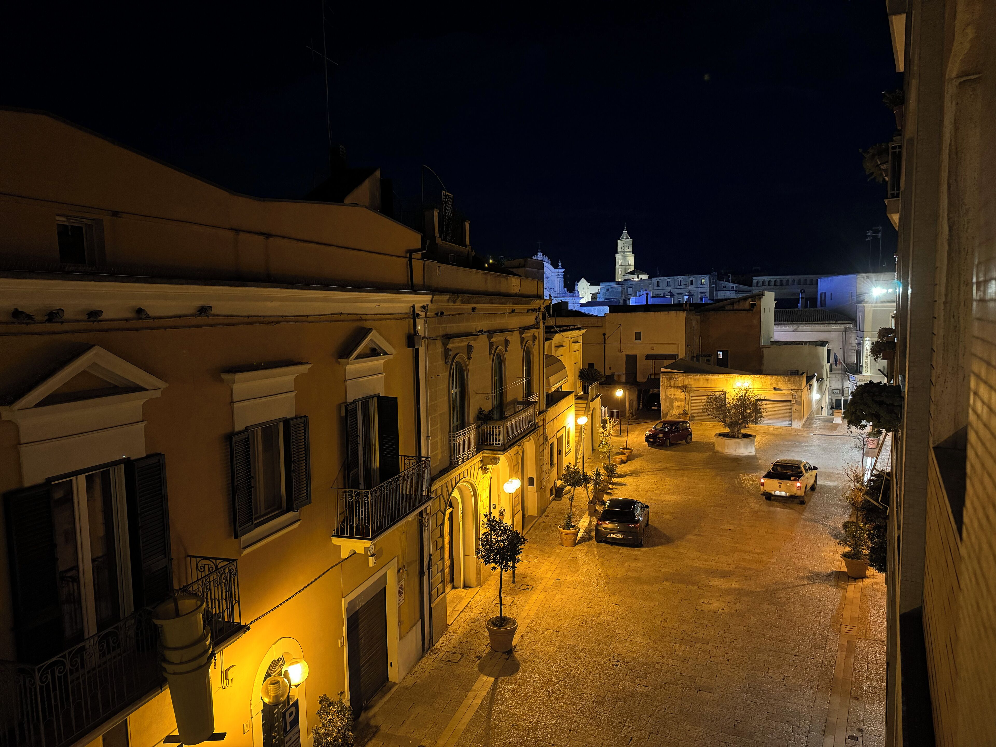 A nighttime view of a narrow street lined with buildings, featuring a parked car and a few potted plants.