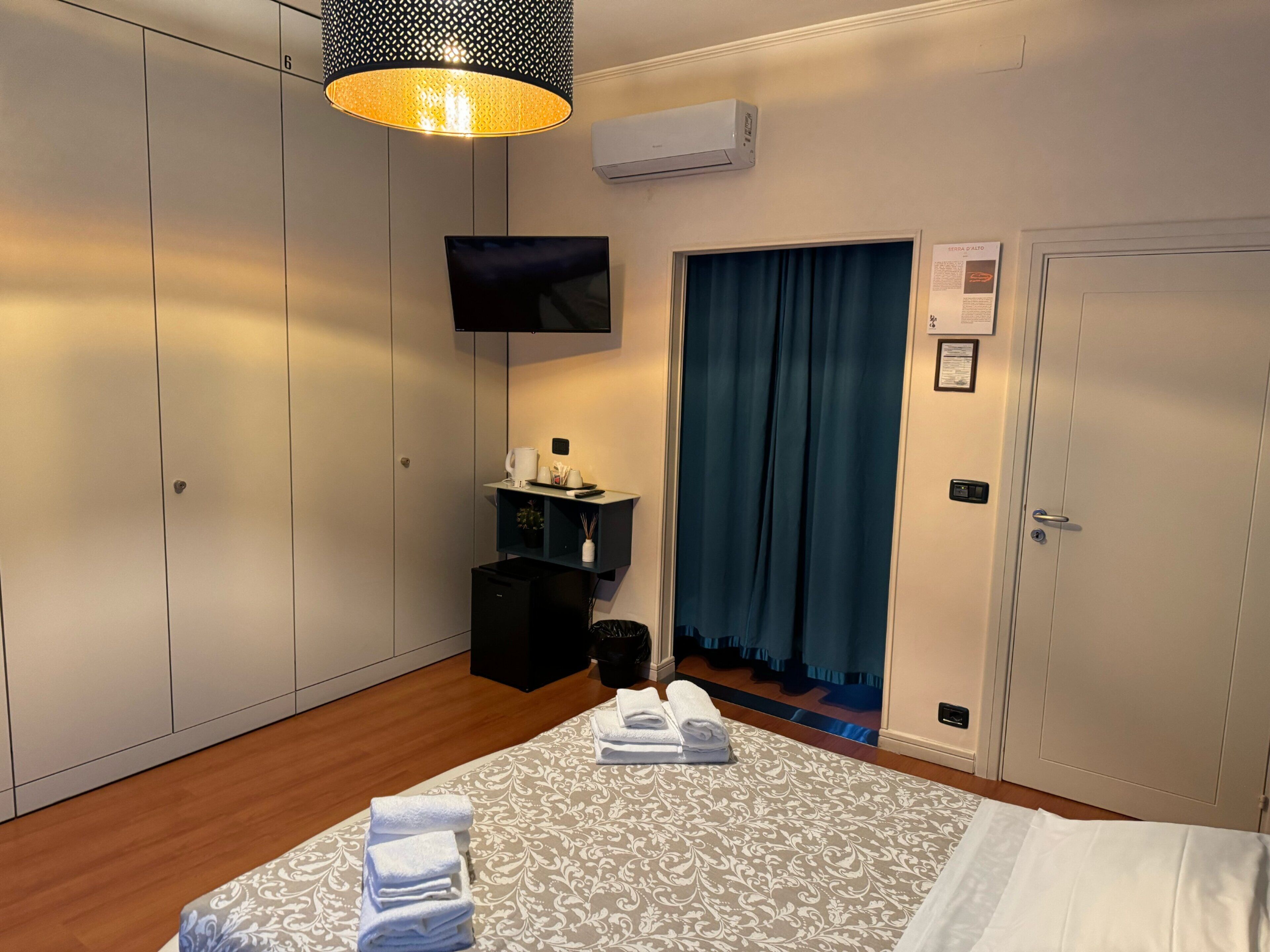 Une chambre d’hôtel équipée d’un lit, d’une télévision, d’un bureau avec un ordinateur, d’une armoire et d’une salle de bain visible à travers un rideau.
