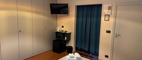 A hotel room with a bed, a TV, a desk with a computer, a wardrobe, and a bathroom visible through a curtain.