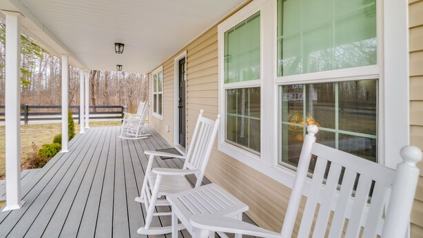 Eine überdachte Veranda mit weißen Schaukelstühlen und einem Tisch, beige Farbe an der Fassade und großen Fenstern.