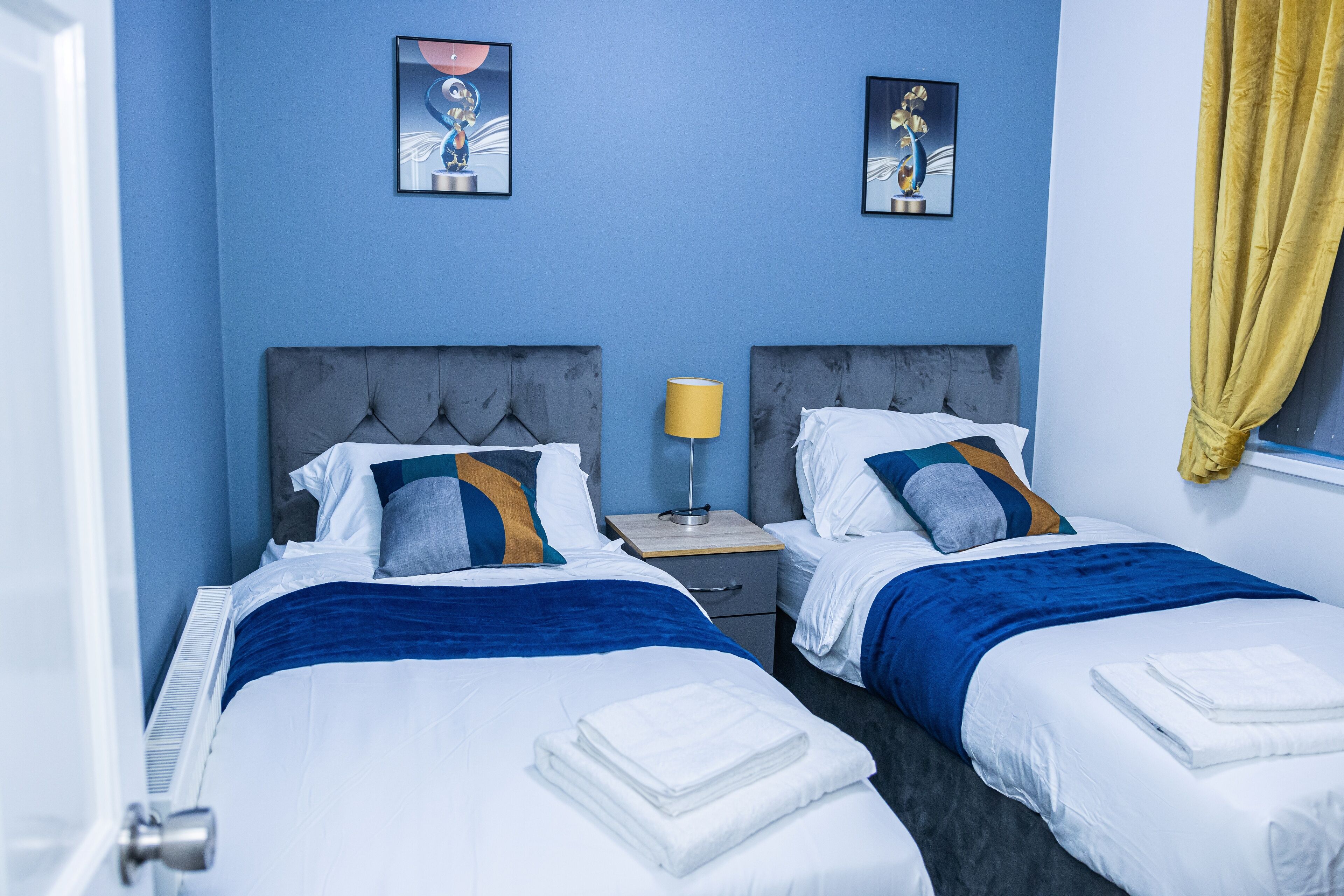 A hotel room with two beds, a nightstand, and a window with yellow curtains.