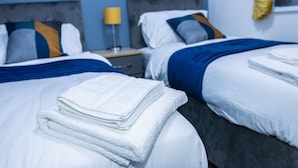 A hotel room with two beds, each with white bedding and blue accents, a nightstand with a lamp, and a window with yellow curtains. - Impeccable 4-bed House in Telford, West Midlands (Telford)