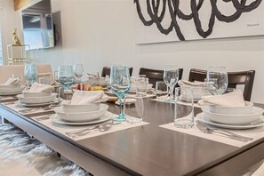 Uma mesa de jantar posta com pratos brancos, copos transparentes e talheres, com um quadro abstrato moderno na parede ao fundo.