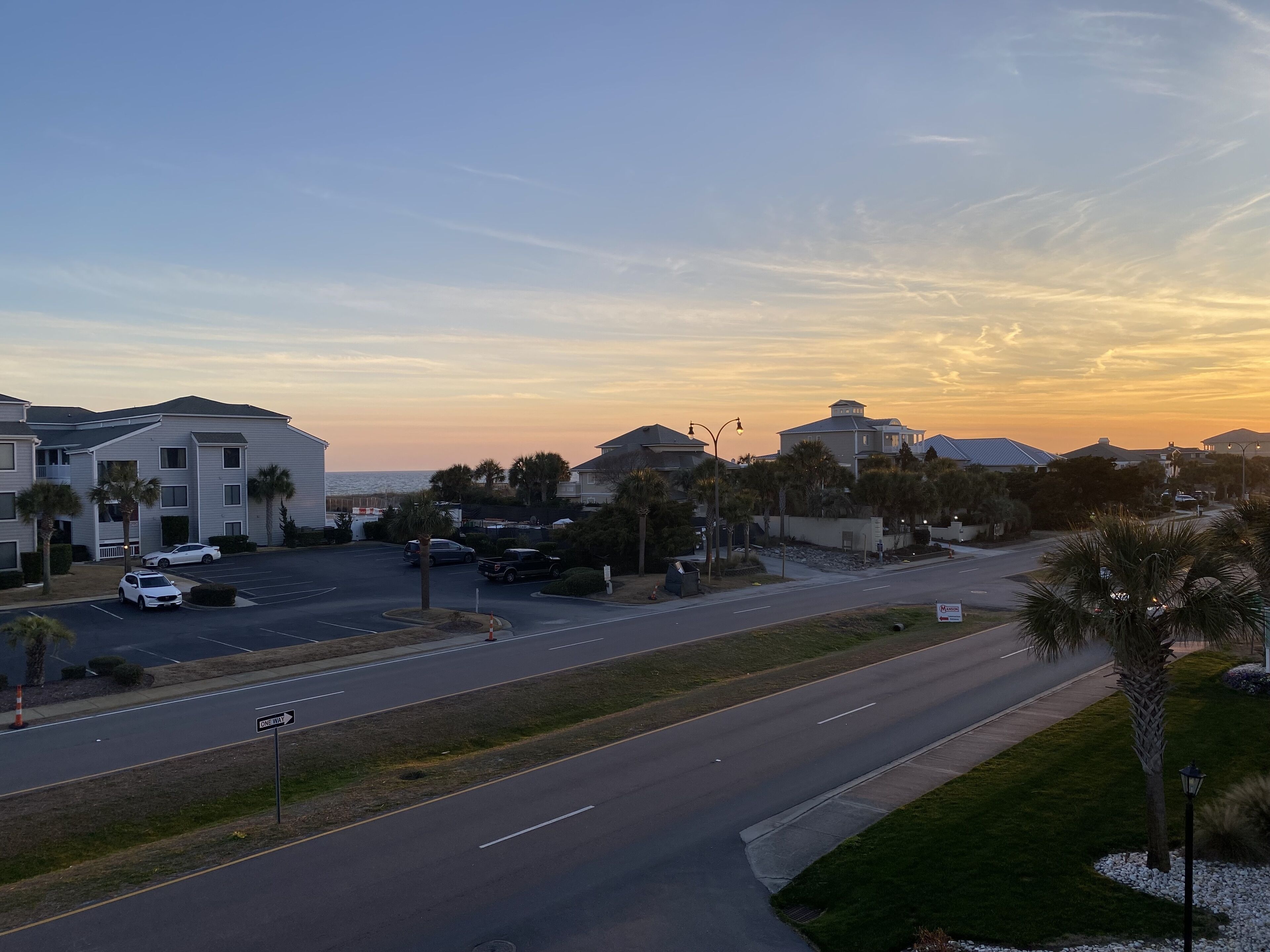 A residential area with a road, parked cars, and buildings with a view of the ocean at sunset.