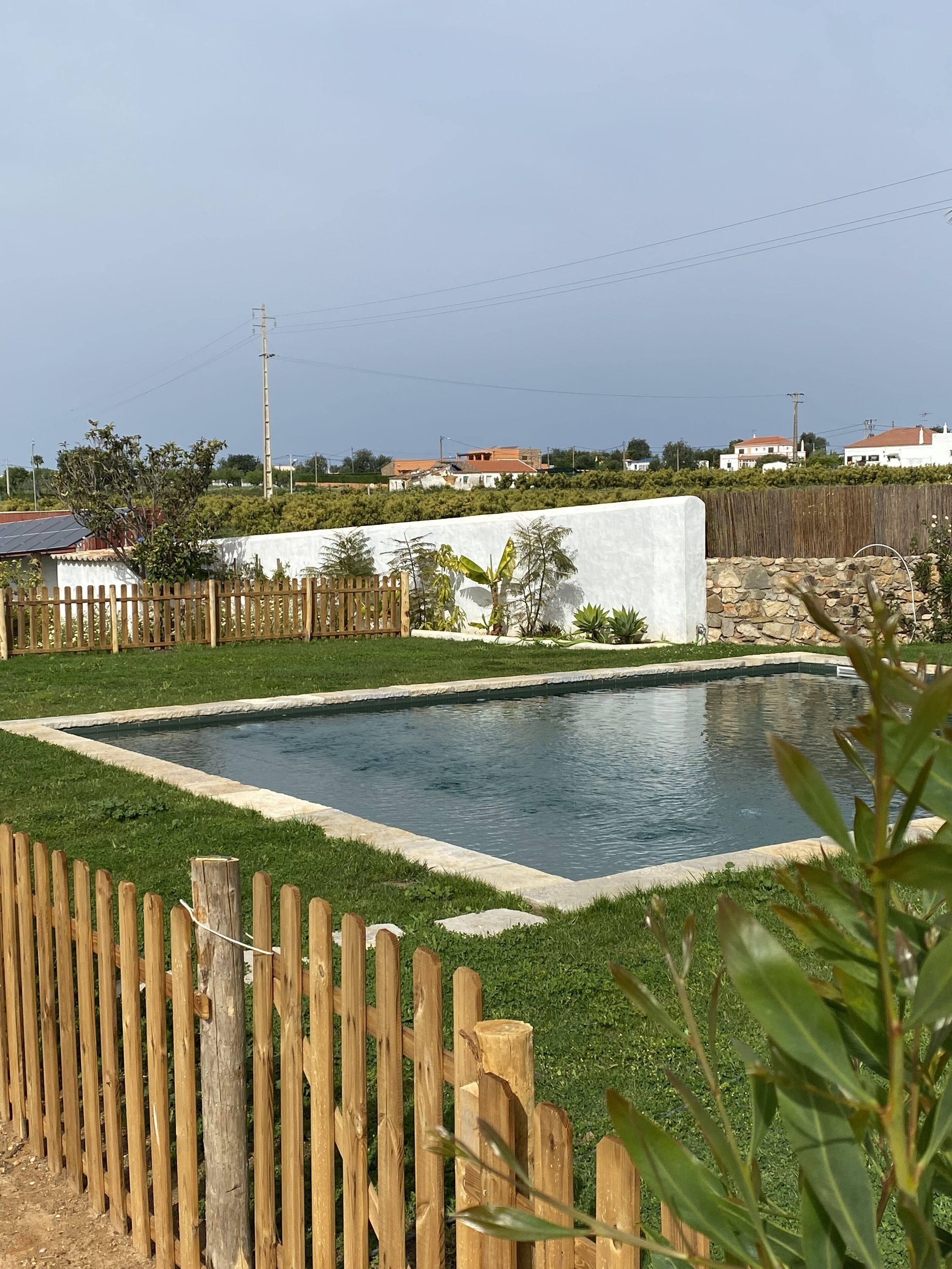 Une piscine rectangulaire bordée de pierres, entourée d’une clôture en bois et d’une végétation luxuriante.