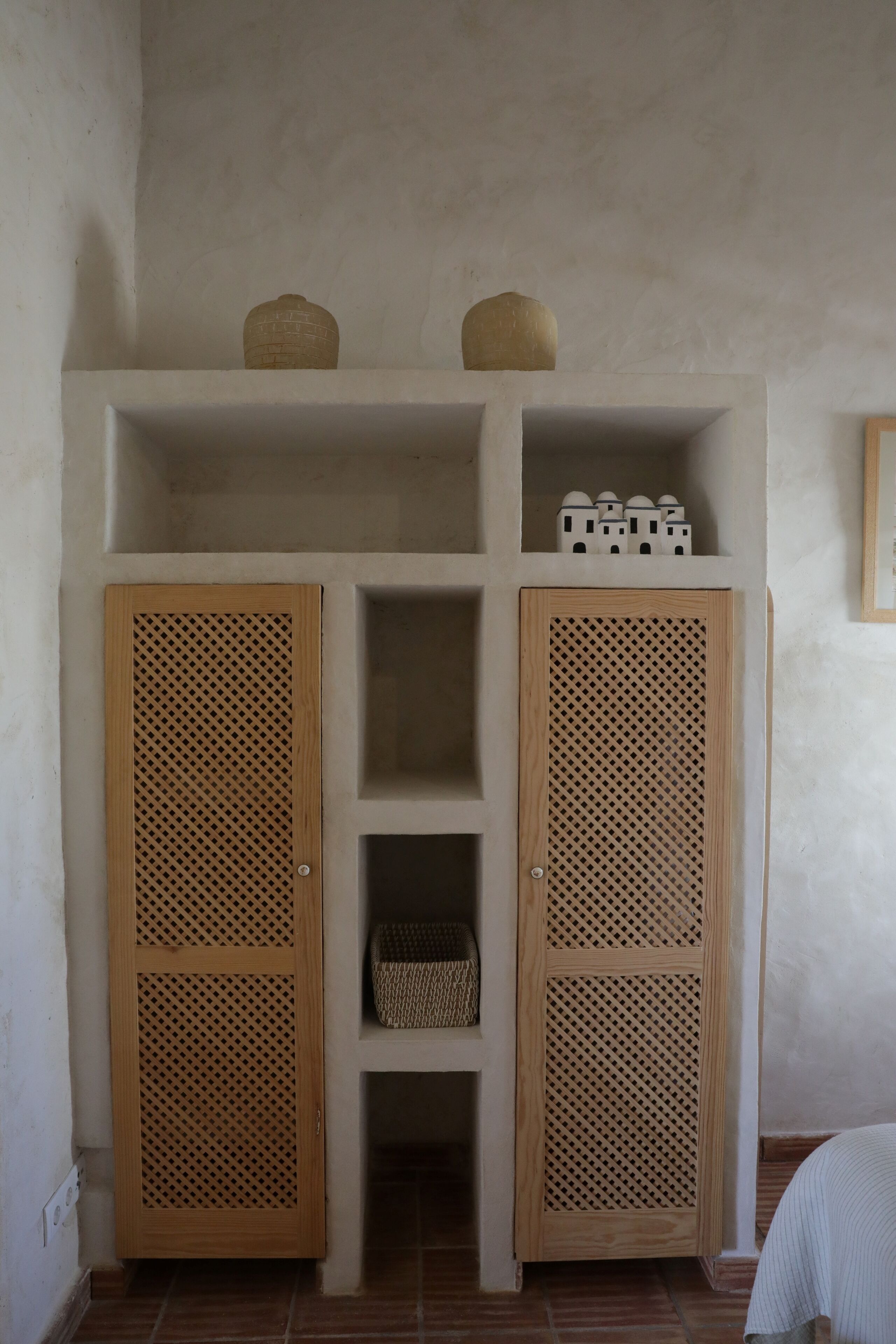 A room with two wooden louvered doors, a shelf with baskets, and a small white bed.