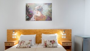 A hotel room with a wooden headboard, two white pillows, a bed with a beige bedspread, a small wooden nightstand, a glass-top desk, and a painting on the wall. - Room-comfort Double or Twin Room or Semi (Paros)
