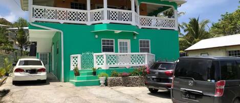 A two-story building with a green facade, white balconies, and a front porch with potted plants.