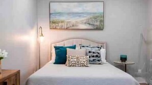 Een slaapkamer met een bed, een schilderij van een strand, een nachtkastje en een bedtafel.