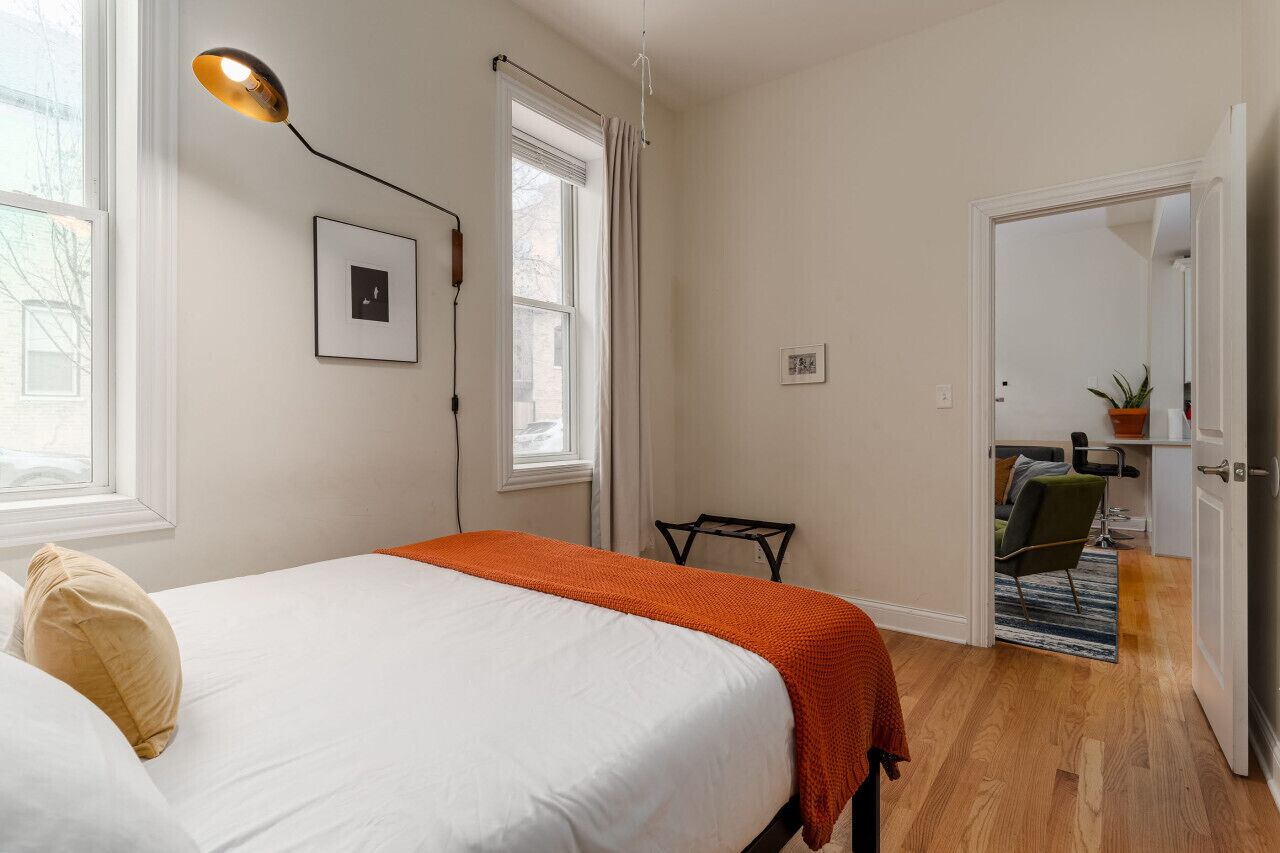 Ein Schlafzimmer mit Bett, Nachttischlampe, Fenster, Tür und einem kleinen Tisch mit Stuhl.