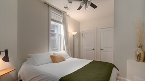 A bedroom with a bed, a ceiling fan, a window with curtains, a bedside lamp, and a wall-mounted vase. - Flexhome East Village • Modern 2BR • Quiet Street (Chicago)