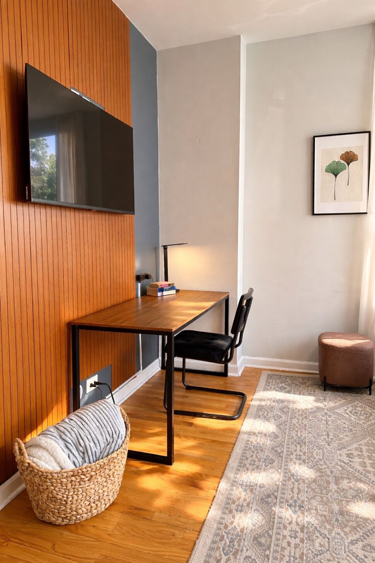 Une pièce moderne avec un mur lambrissé en bois, un téléviseur à écran plat fixé au-dessus d’une table à manger en bois, une chaise noire, un pouf en fibres tressées et un tapis à motifs.