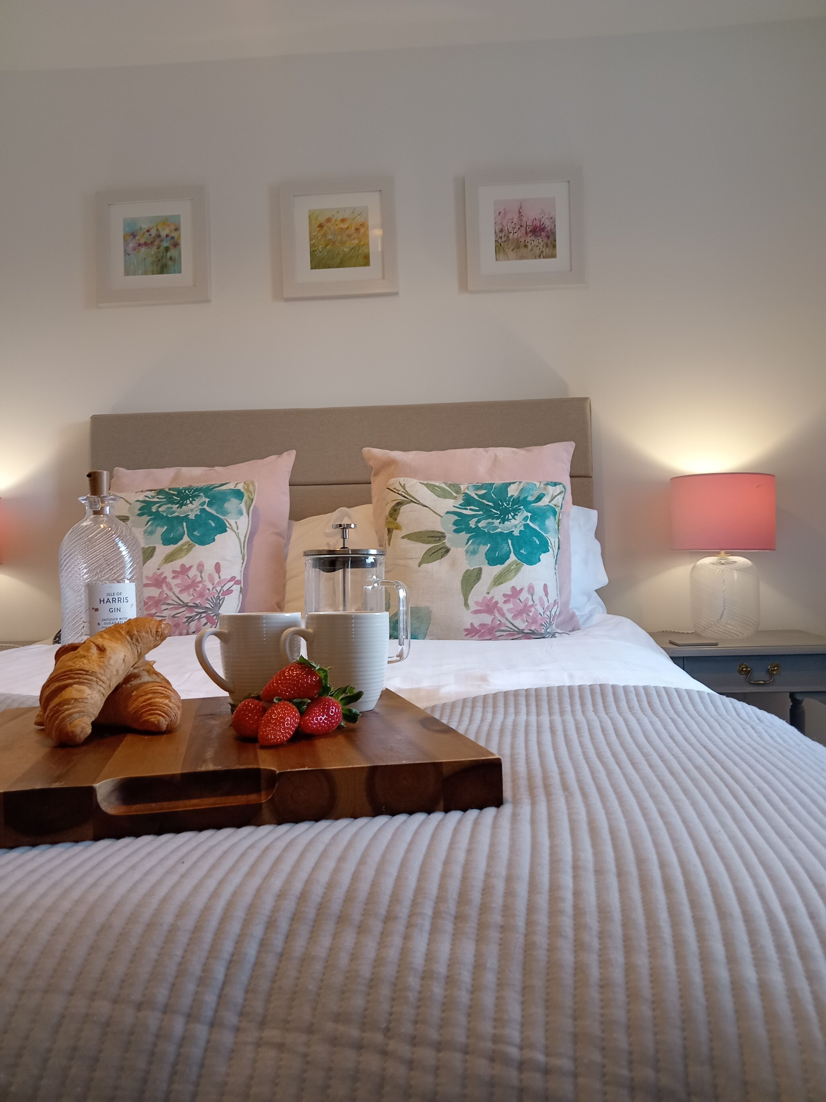 A neatly made bed with a white duvet, a wooden tray with croissants, strawberries, a mug, and a bottle of gin.