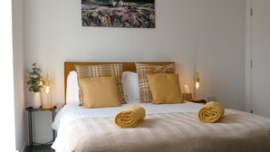 A neatly made bed with a patterned bedspread and two rolled towels, a painting on the wall, and a bedside lamp.