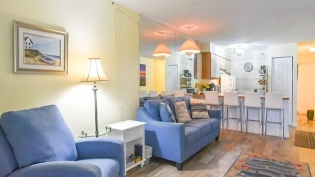Een woonkamer met een blauwe bank, een kleine tafel en een lamp. Er is een keuken met een bar en stoelen.
