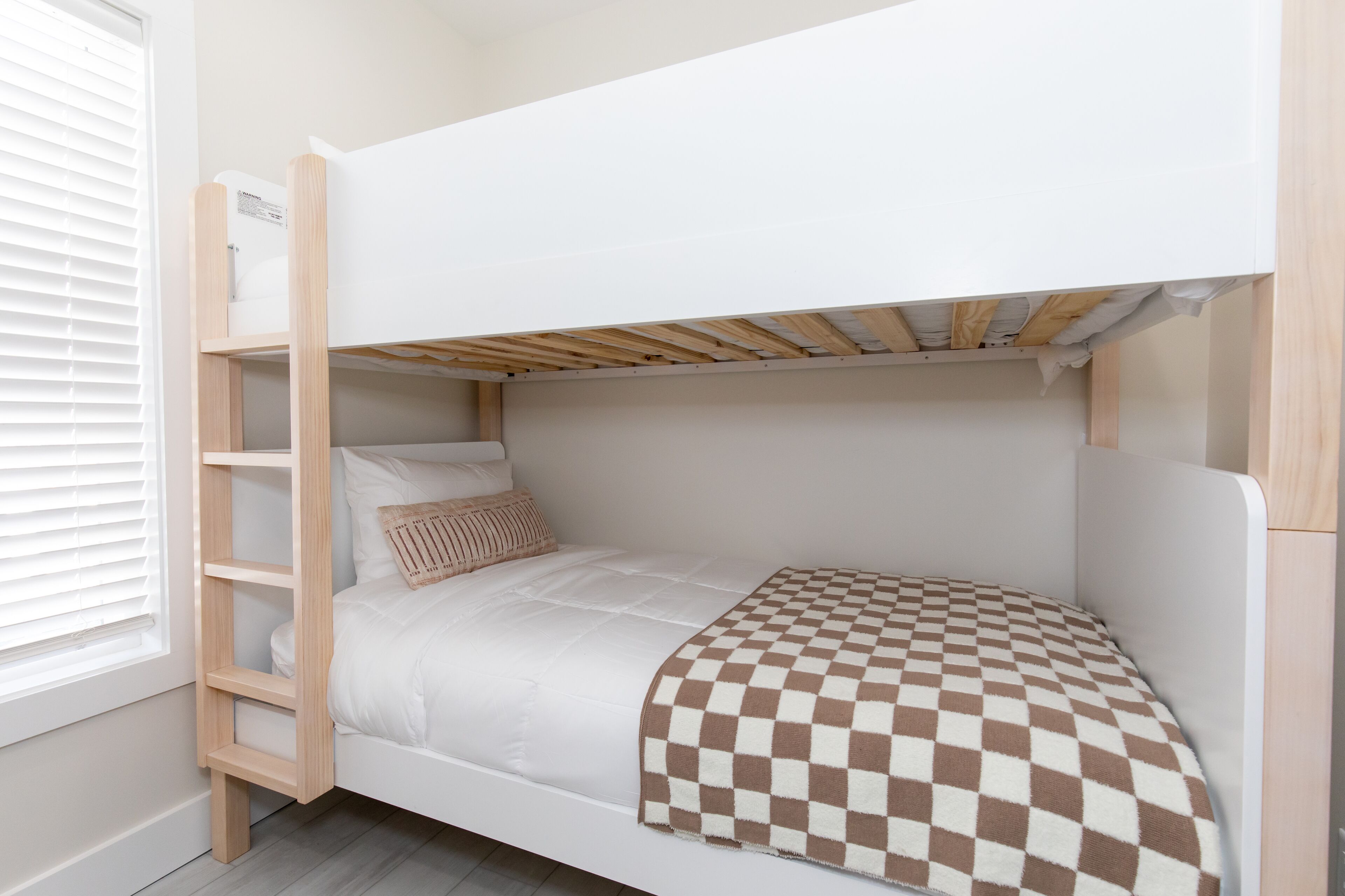 A bunk bed with a checkered blanket, a wooden ladder, and a window with blinds.