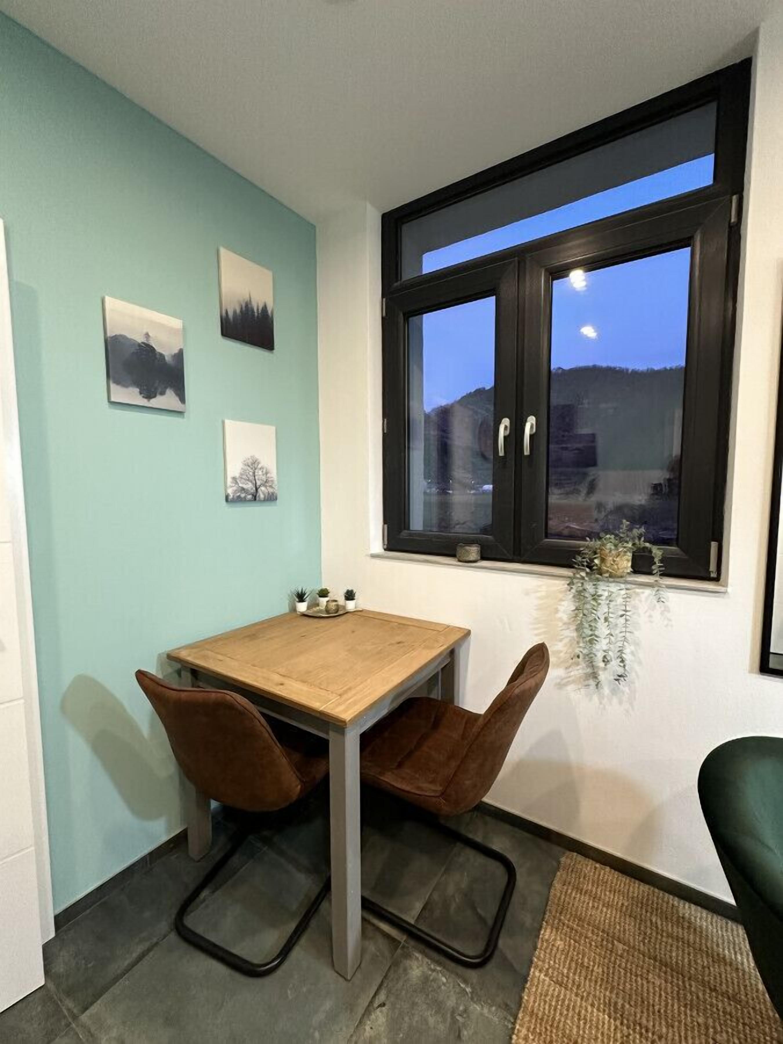 A dining area with a wooden table and two brown chairs, a window with a view of the night sky, and three framed pictures on the wall.