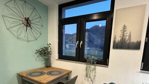 A dining area with a wooden table, chairs, a decorative wall clock, a window with a view of mountains, and a blue chair. - Relaxen im Weindorf Mayschoß Fewo 3 (Mayschoß)