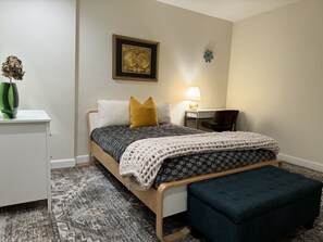 A neatly arranged bedroom with a bed, bedside table, lamp, chair, and a decorative item on the wall.