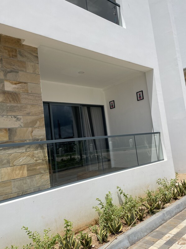 A modern building with a stone facade, a balcony with glass railings, and a small garden area with plants.