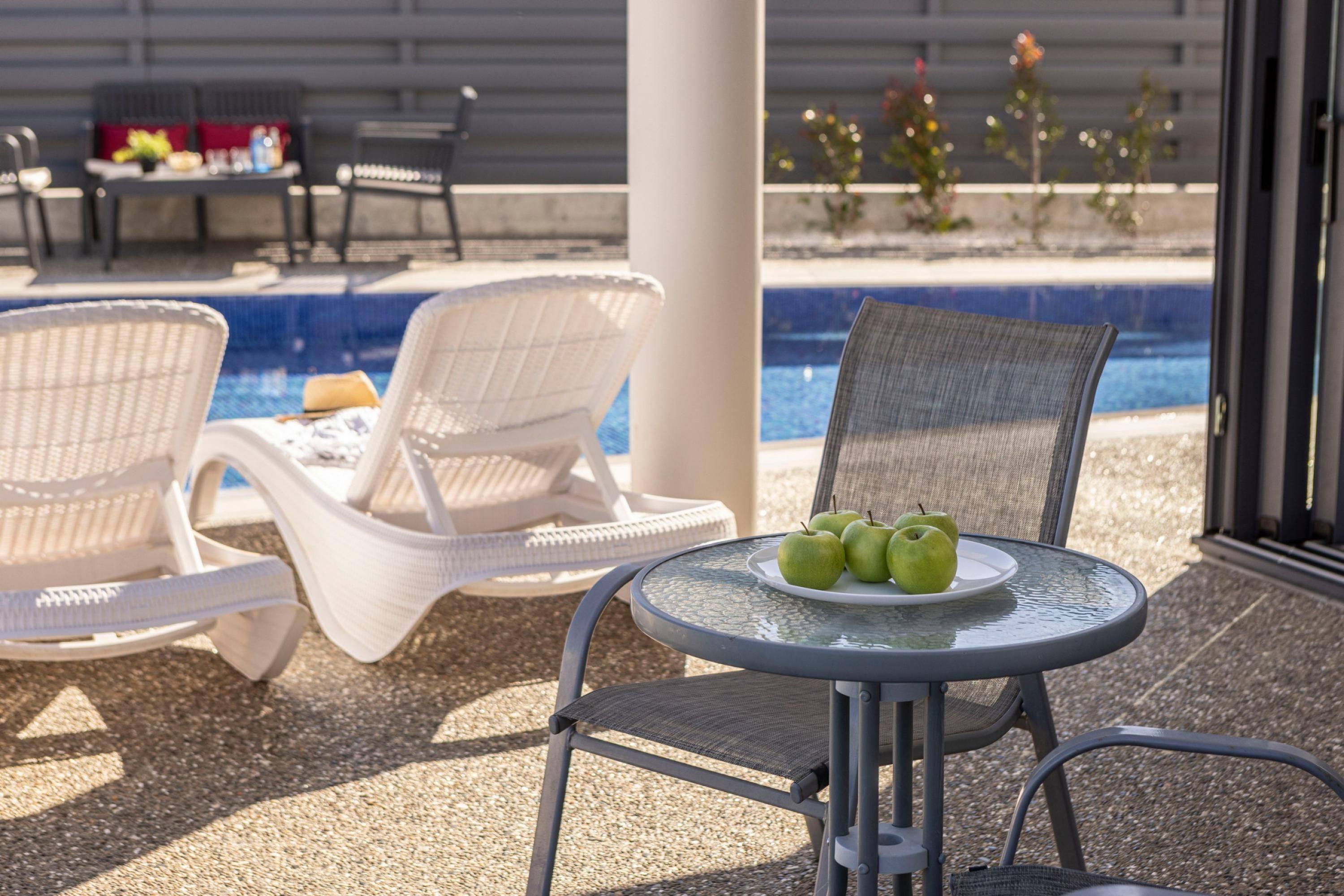 A poolside area with wicker lounge chairs, a round table with a plate of green apples, and a view of the pool and plants in the background.