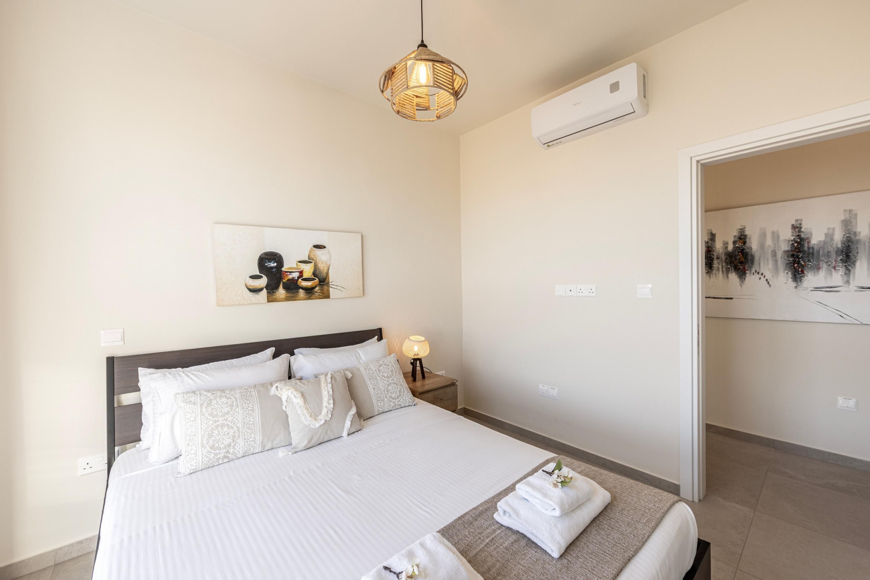 Ett välordnat hotellrum med en stor säng, ett sängbord med en lampa, en väggmonterad luftkonditioneringsanläggning och ett inramat konstverk som föreställer en stadsbild.