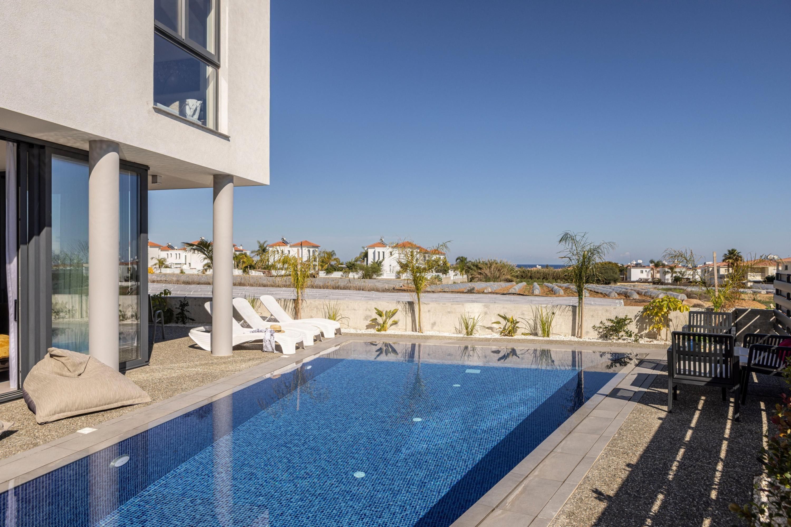 A modern pool area with clear blue water, white lounge chairs, and a view of the sea and buildings in the background.