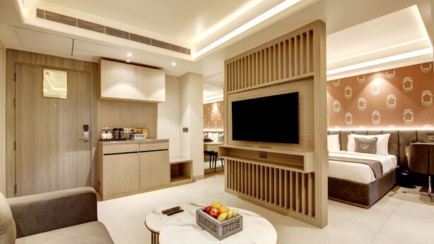 A modern hotel room with a flat-screen TV, a dining area, a sofa, a small table with fruit, and a bed with a headboard. - Stepstones Hotels and Inn (Gurugram)